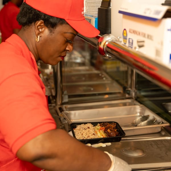 A Peppas Jerk Chicken employee in a red uniform preparing a takeout container of jerk chicken and rice.