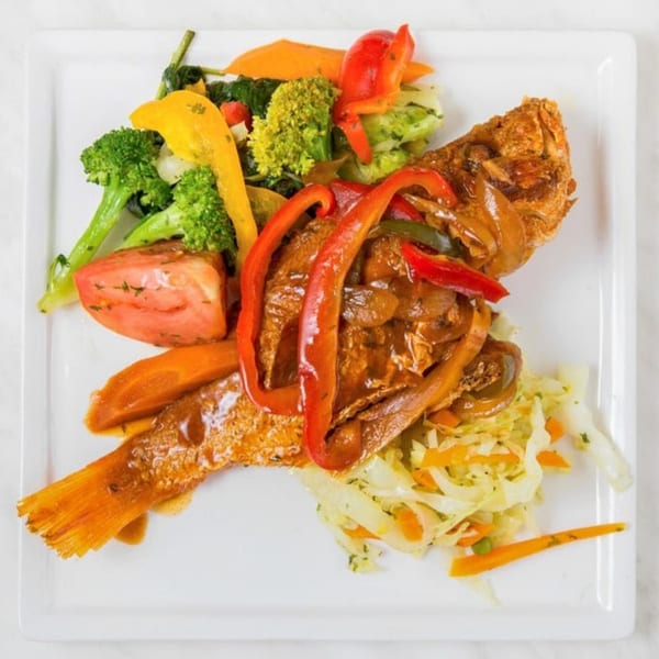 Whole fried fish with brown sauce, red peppers, and a side of mixed vegetables on a white plate.