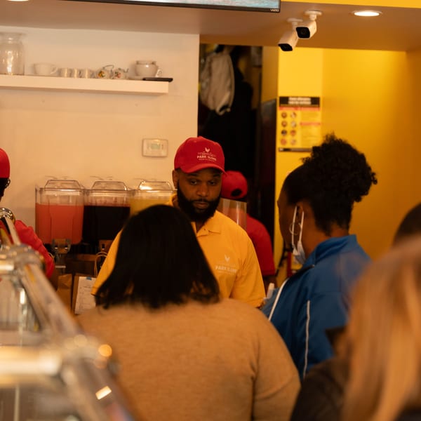 A Peppas Jerk Chicken employee in a red hat and yellow shirt serves customers at the counter.