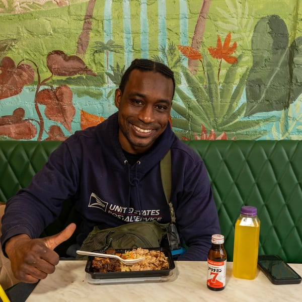 A smiling man in a USPS hoodie gives a thumbs-up while sitting with a meal at Peppas Jerk Chicken.