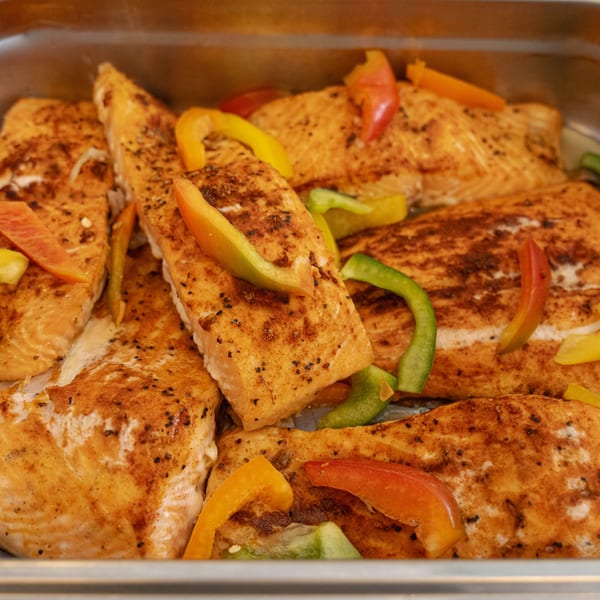 Close-up of seasoned salmon fillets with colorful bell pepper strips in a metal tray.