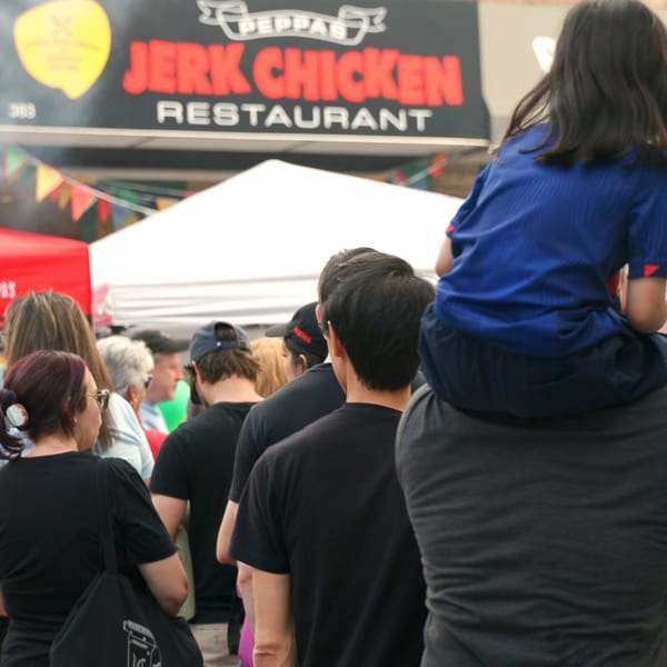 A crowd of people, including a child on shoulders, waiting in line at Peppas Jerk Chicken.